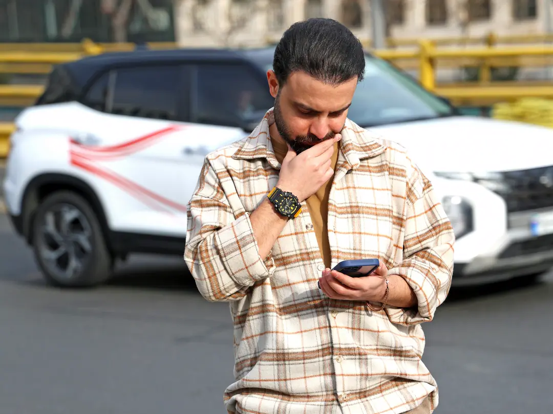 Homem consultando o celular em pé na frente de um carro branco, antes de fechar negócio.